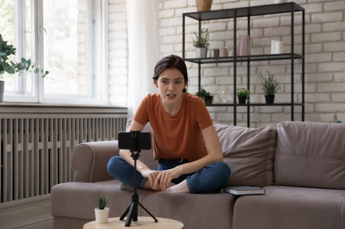 Young female vlogger sits down on couch, in front of cell phone on a tripod to make a video for social media