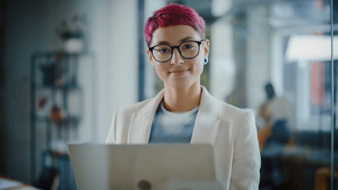 Modern Office: Portrait of Beautiful Authentic Specialist with Short Pink Hair Standing, Holding Laptop Computer, Looking at Camera, Smiling Charmingly. Working on Design