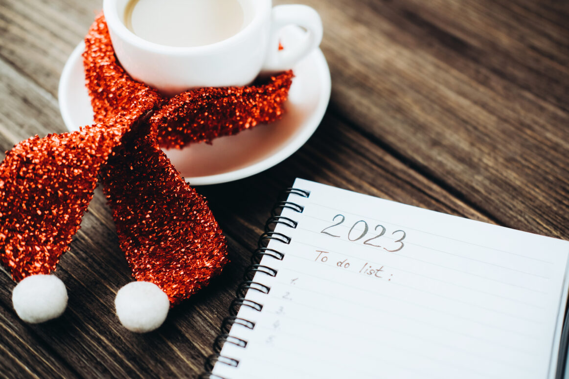 2023 To do list written on notepad close to white glossy coffee cup wrapped in Santa Claus scarf in saucer on dark wooden surface.