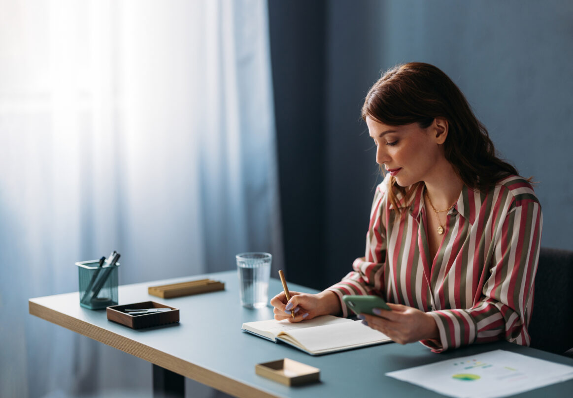 A businesswoman is attentively writing in a notebook, with a smartphone in hand, at her modern office desk, embodying productivity and organization.