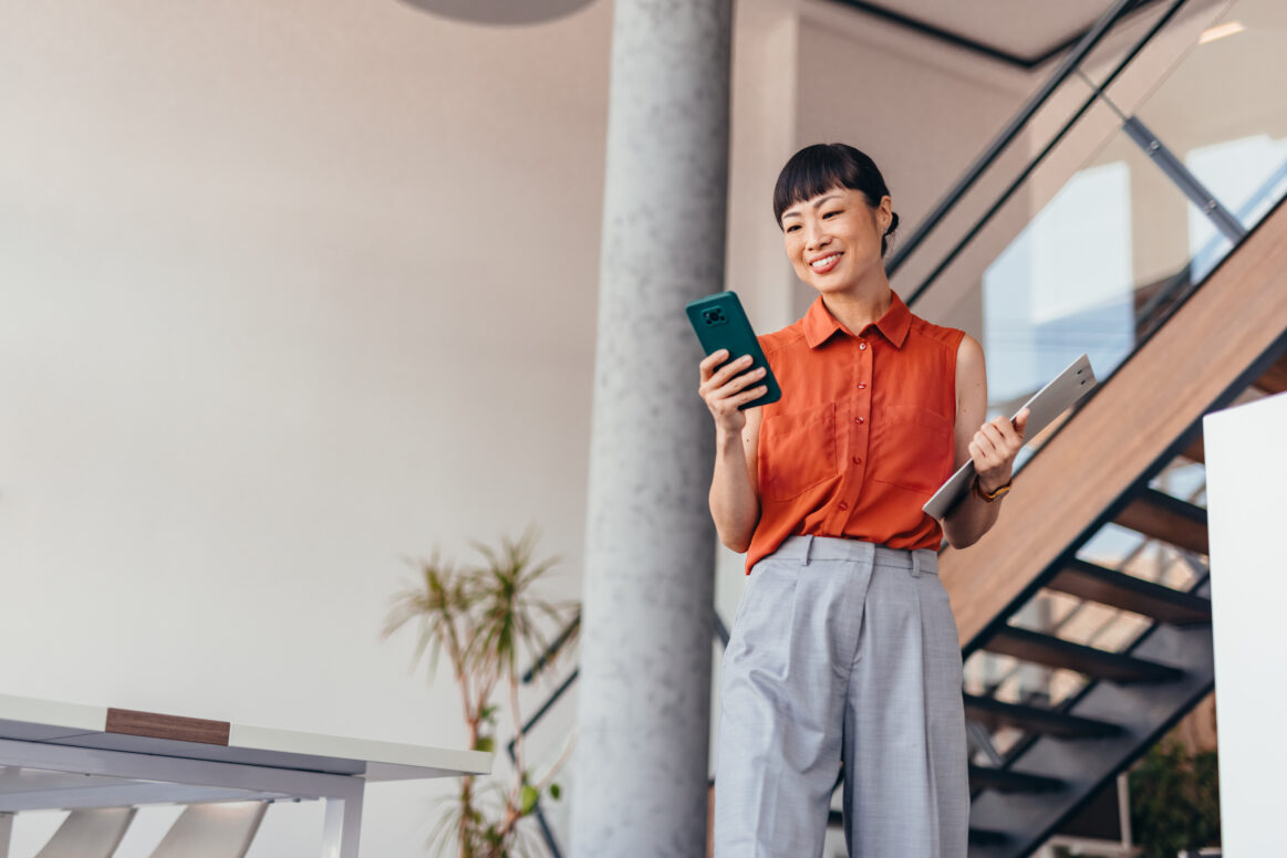 A professional Asian woman in an office reviews documents on a clipboard while using a mobile phone, symbolizing multitasking and communication in a modern work environment.