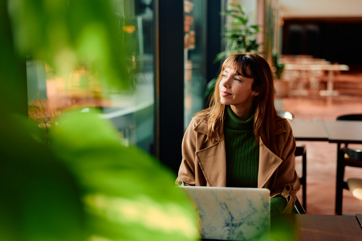 Woman freelancer taking a break from work, sitting at a cafe table with her laptop, contemplating while looking away with sunlight streaming through the window